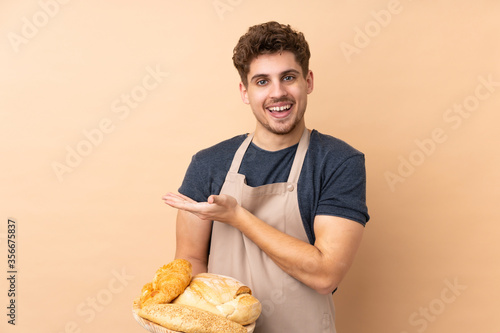 Male baker holding a table with several breads isolated on beige background extending hands to the side for inviting to come