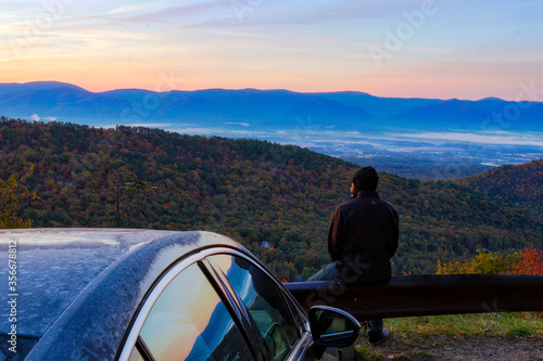 Фотография Man parks car at a mountain lookout and sits on guardrail to enjoy the sunrise on a chilly autumn morning taking in the valley below and the mountains in the distance