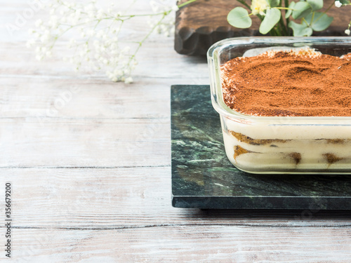 Traditional coffee tiramisu dessert with mascarpone cheese and cocoa powder in glass container on green marble board
