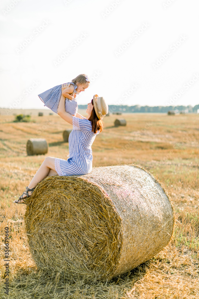 Young charming mother with her adorable baby girl, wearing in similar striped blue dresses, sitting on a sheaf of straw in a beautiful field and playing together. Mom holds her baby on hands Young charming mother with her adorable baby girl, wearing in similar striped blue dresses, sitting on a sheaf of straw in a beautiful field and playing together. Mom holds her baby on hands
