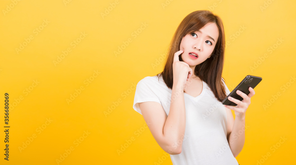 Asian happy portrait beautiful cute young woman teen standing wear t-shirt her using holding smart mobile phone and thinking looking to side isolated, studio shot yellow background with copy space Asian happy portrait beautiful cute young woman teen standing wear t-shirt her using holding smart mobile phone and thinking looking to side isolated, studio shot yellow background with copy space