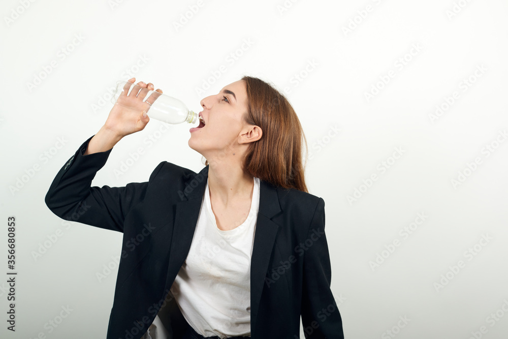 Drinking water from plastic bottles, quenches thirst after fitness indoors. Lose weight. Healthy lifestyle. Young attractive woman with brown hair in a light t-shirt, black jacket on white background Drinking water from plastic bottles, quenches thirst after fitness indoors. Lose weight. Healthy lifestyle. Young attractive woman with brown hair in a light t-shirt, black jacket on white background