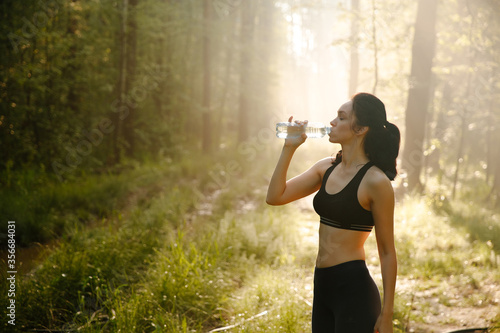 The girl is engaged in sports in the morning, during rest drinks cool water in a pine forest green and lush beautiful. Sporty girl and the concept of lifestyle.