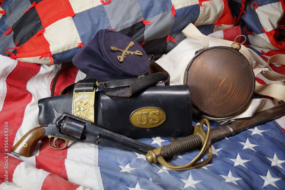 Obraz premium Artifacts from the American Civil War. Union army hat gear sit on top of an American flag and a patriotic quilt. A pistol and saber from the US Cavalry, canteen and leather items make up the display.