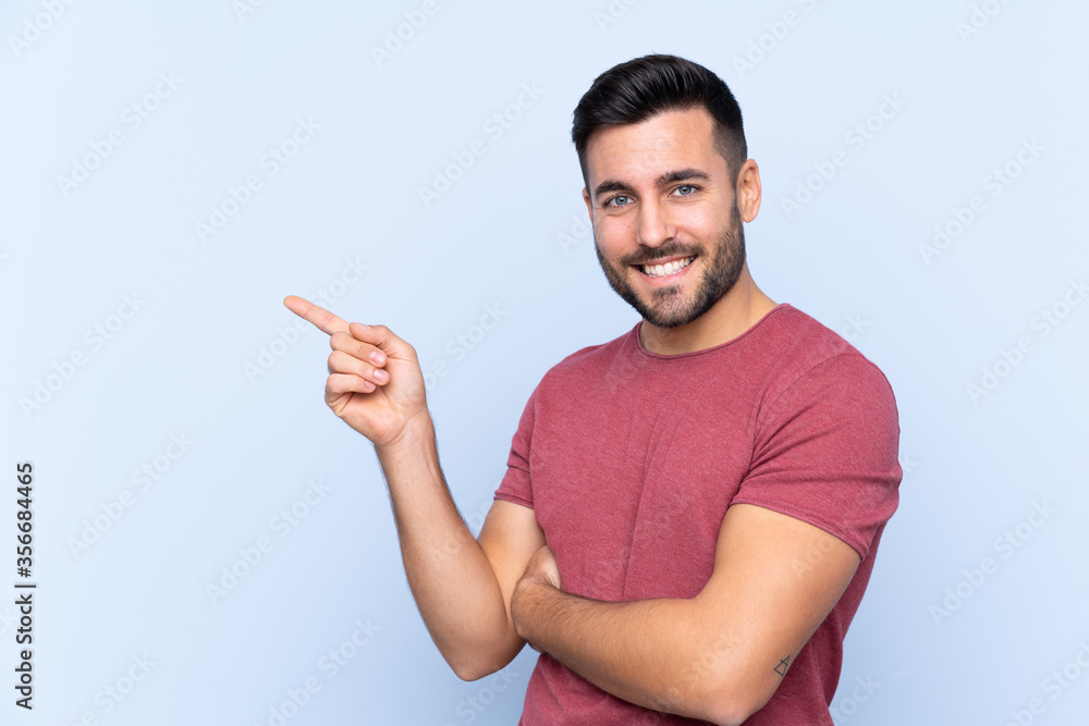 Young handsome man with beard over isolated blue background pointing finger to the side