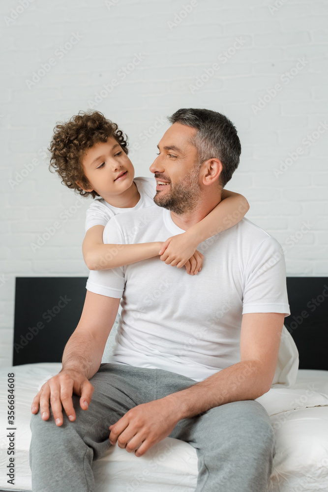 curly son hugging happy bearded father and looking at each other in bedroom curly son hugging happy bearded father and looking at each other in bedroom
