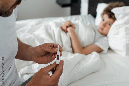 selective focus of father holding digital thermometer near sick son in bed