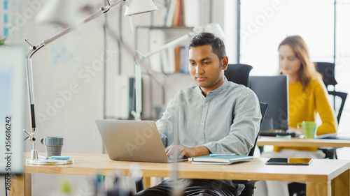 Smart and Handsome Indian Information Technology Specialist Sitting at His Desk works on a Laptop. In the Background Modern Office with Diverse Team of Young Professionals Working.