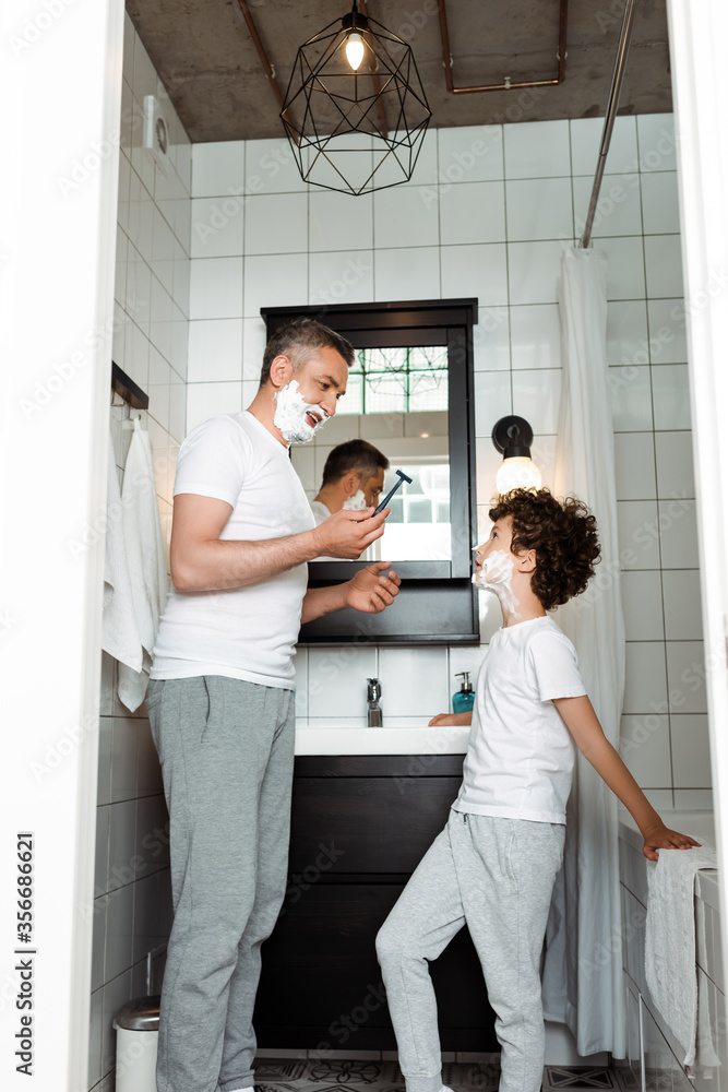 Obraz premium handsome father with shaving foam on face holding razor near curly son