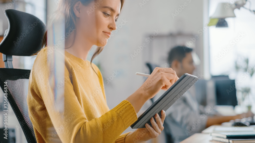 Talented Female Designer Sitting at Her Desk She's Drawing, Writing and Using Pen with Digital Tablet Computer. Focus on Hands with Pen. Bright Office where Diverse Team of Young Professionals Work.