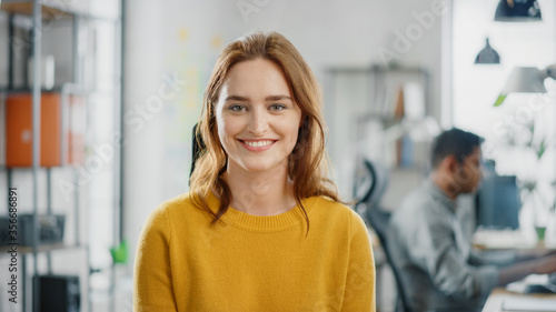 Portrait of Beautiful Young Woman with Red Hair Wearing Yellow Sweater Smiling at Camera Charmingly. Successful Woman Working in Bright Diverse Office. photo