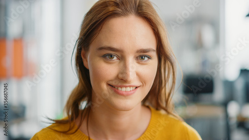 Portrait of Beautiful Young Woman with Red Hair Wearing Yellow Sweater Looking Up to the Camera and Smiling Charmingly. Successful Woman Working in Bright Diverse Office.