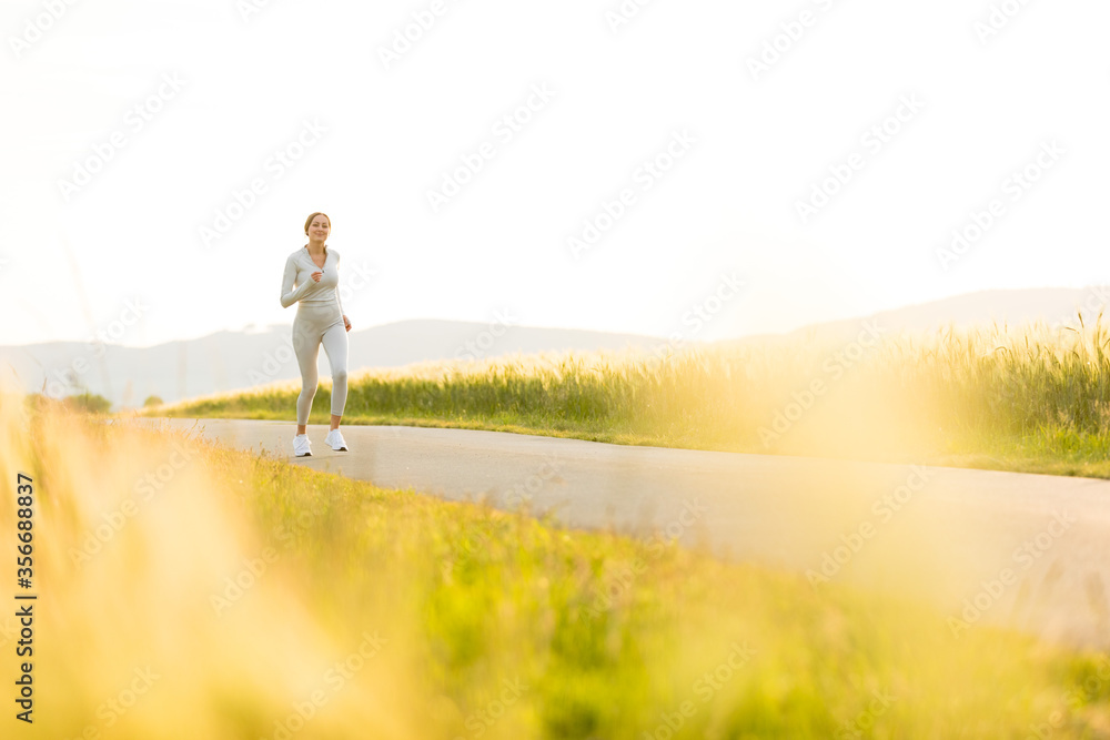 Sportliche junge Frau joggt abends zwischen Feldern Sportliche junge Frau joggt abends zwischen Feldern