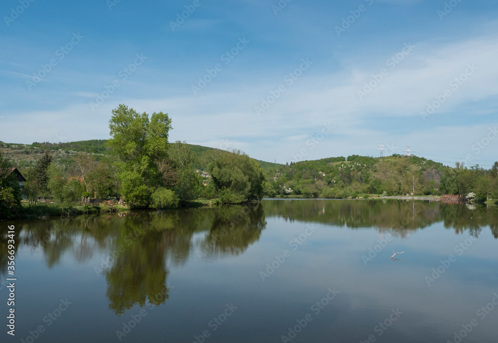 view on river Berounka from pedestrial bridge from village Zadni Treban to Hlasna treban in central Bohemian region, green lush trees reflecting in water, blue sky, spring sunny day