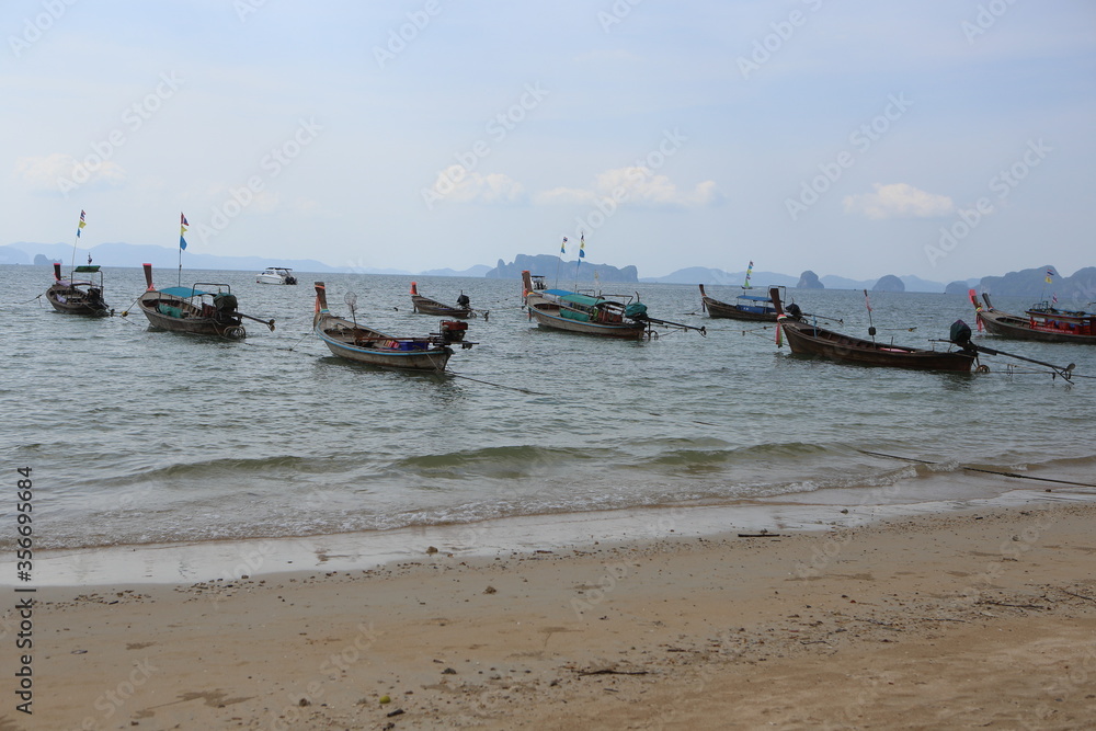 beautiful day shot of rows of long boats in shallow ocean waters by the shore of yellow sand beach with islands, hills and mountains, blue sky and white clouds in the background. Krabi, Thailand beautiful day shot of rows of long boats in shallow ocean waters by the shore of yellow sand beach with islands, hills and mountains, blue sky and white clouds in the background. Krabi, Thailand