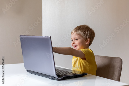 A cute little boy with blond hair sits at a table with a laptop and waves his hand. The boy smiles. He's got virtual communication.
