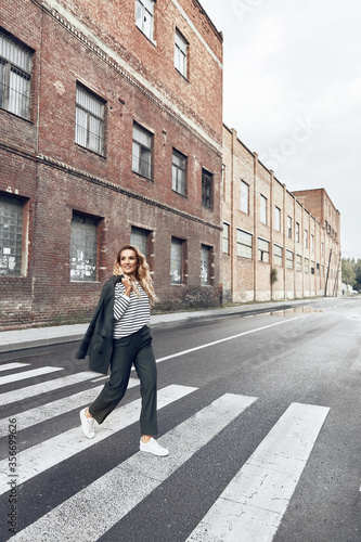 streetstyle photo of woman with long blond curly hair сrosses the street, looks to the left and smiles, she is wearing black pants, striped blouse and white sneakers, she is holding a jacket © monchak
