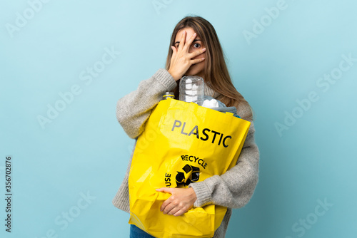 holding a bag full of plastic bottles to recycle over isolated blue covering eyes and looking through fingers
