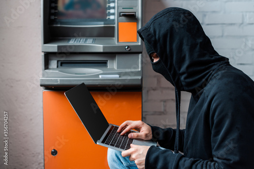 Side view of hacker in mask using laptop with blank screen near atm photo