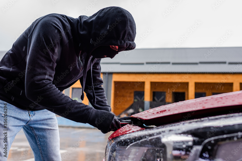Side view of robber in balaclava holding screwdriver while opening car hood on urban street Side view of robber in balaclava holding screwdriver while opening car hood on urban street