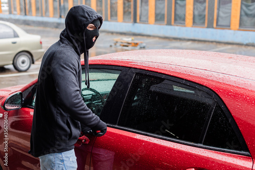 Robber in balaclava and leather gloves looking away while opening car with screwdriver on urban street