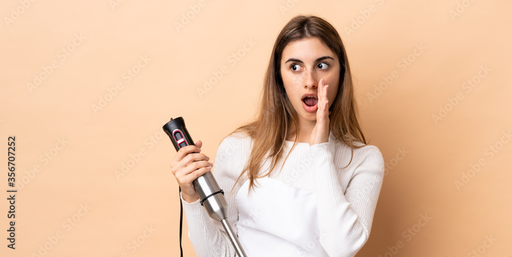 Woman using hand blender over isolated background whispering something with surprise gesture while looking to the side Woman using hand blender over isolated background whispering something with surprise gesture while looking to the side