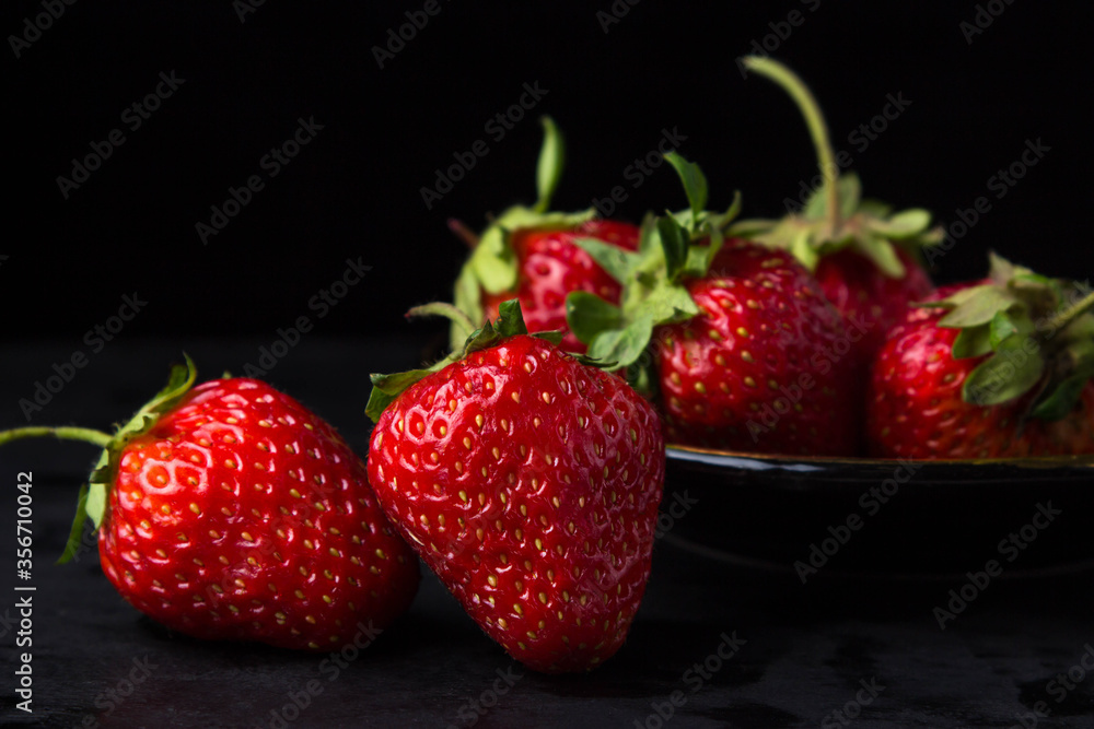 Strawberries on a black background. Two strawberries in the foreground. behind a plate with strawberries. Creative photo of strawberries Strawberries on a black background. Two strawberries in the foreground. behind a plate with strawberries. Creative photo of strawberries