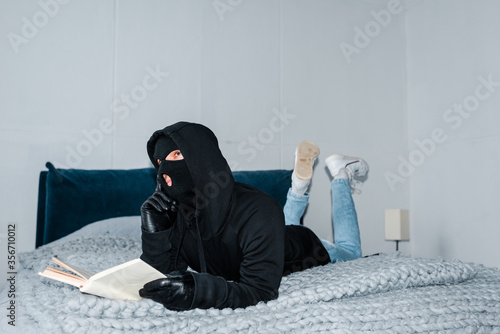 Photo Pensive robber in balaclava holding book while lying on bed