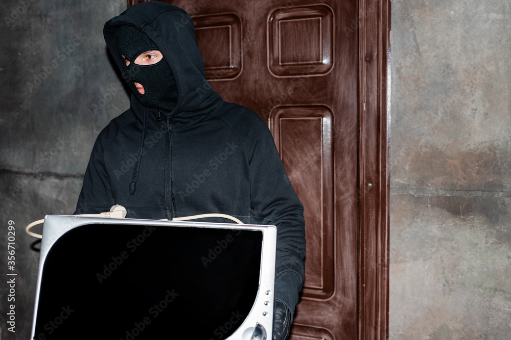 Robber in balaclava holding microwave oven during house stealing