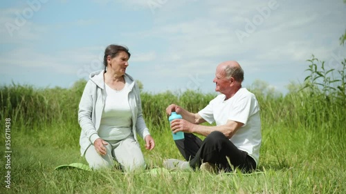 caring elderly man gives cool clean water in sports nutrition bottles to his dear beloved wife after playing sports or meditating, outdoors on background of green grass photo