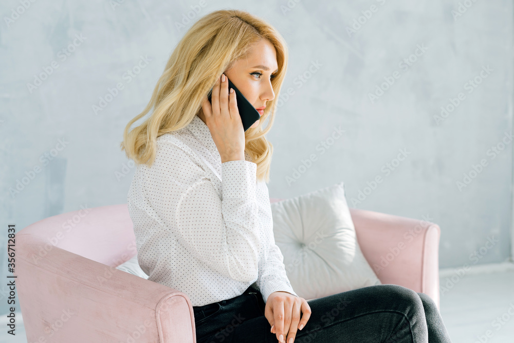 Blonde woman sitting on a pink sofa in a light shirt and sadly talking on the phone