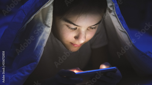 Toned portrait of smiling teenage girl lying in bed with smartphone and browsing internet