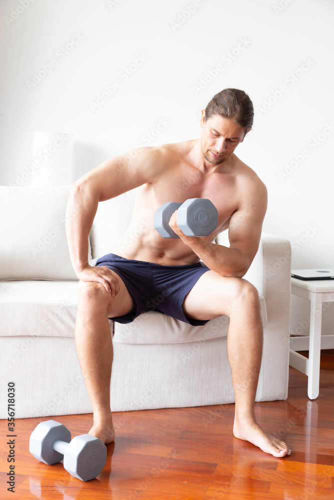 Obraz premium Athletic young man doing exercise with dumbbell in living room at home