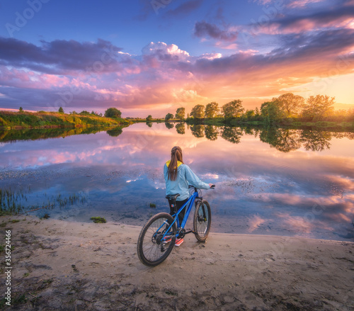 Woman with bicycle on the coast of the river at sunset in summer. Landscape with young girl with mountain bike, sandy beach, purple sky with colorful clouds reflected in water. Sport and travel. Cycle
