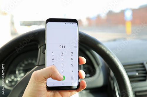 A hand of a car driver holding his phone, dailing the emergency number 911 on the numeric dail pad. A concept of a road accident or aggressive road rage which requieres help of an ambulance or police.