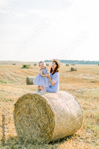 Happy two year old girl child in striped dress playing with young smiling mom in summer autumn field with hay bales, sitting together on hay stack. Summer sunset portrait