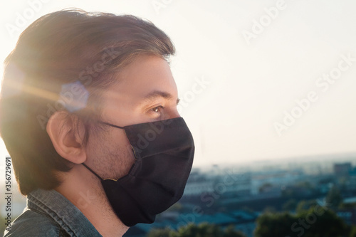 A young man in a black medical mask and shirt stands looking ahead against the blurry backdrop of an industrial city 