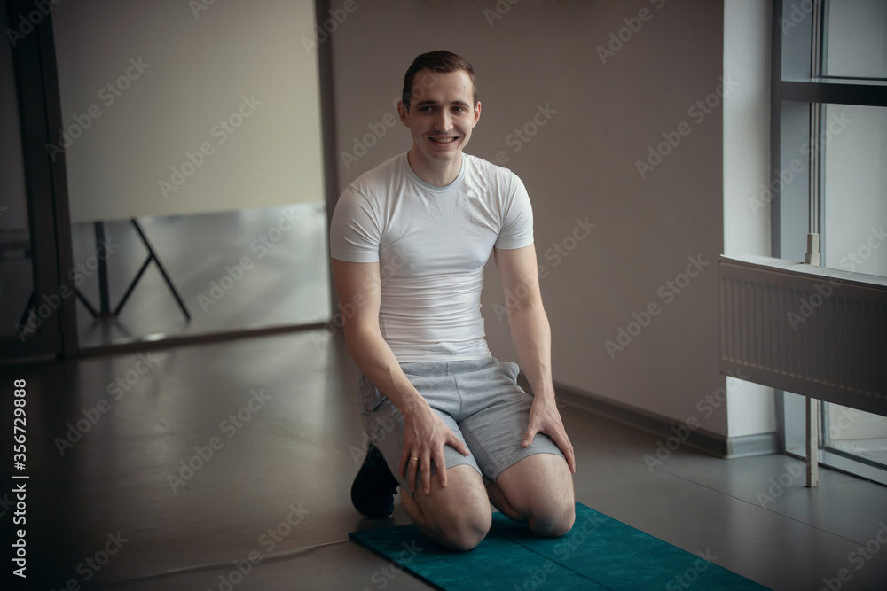 Modern gym - a young man in white t-shirt sitting on the yoga mat Modern gym - a young man in white t-shirt sitting on the yoga mat