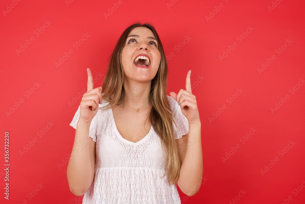 Charming carefree woman with positive expression, points up with both index fingers, dressed in casual clothing, has broad interested smile, isolated over gray background. Look there, please. Charming carefree woman with positive expression, points up with both index fingers, dressed in casual clothing, has broad interested smile, isolated over gray background. Look there, please.