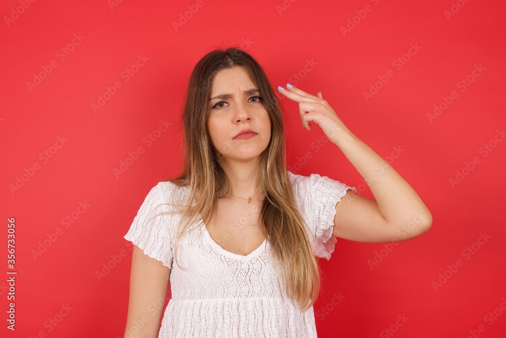 Unhappy blonde young female imitates gun shoot makes suicide gesture keeps two fingers on temples has puzzled expression dressed in casual clothes stands indoors. Unhappy blonde young female imitates gun shoot makes suicide gesture keeps two fingers on temples has puzzled expression dressed in casual clothes stands indoors.