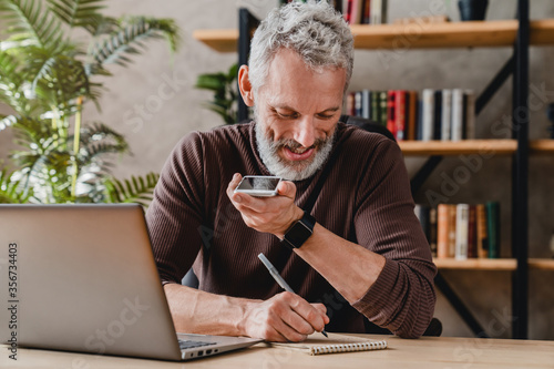 Foto Senior man writing notes in notepad while talking by speakerphone from workspace