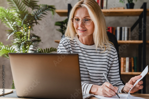 Senior stylish woman taking notes in notebook while using laptop at home office