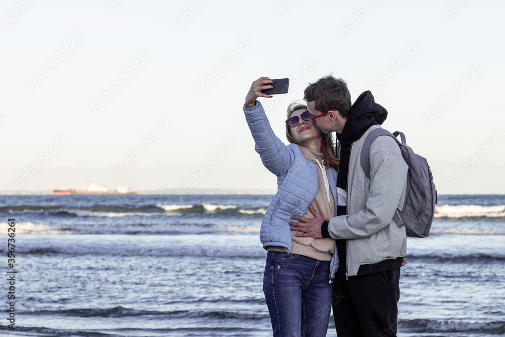 Obraz premium Young couple in love on sea shore taking selfie with mobile phone. Portrait of a romantic couple in love hugging and kissing by the sea. Tourists taking travel photos on their holidays in cold season.