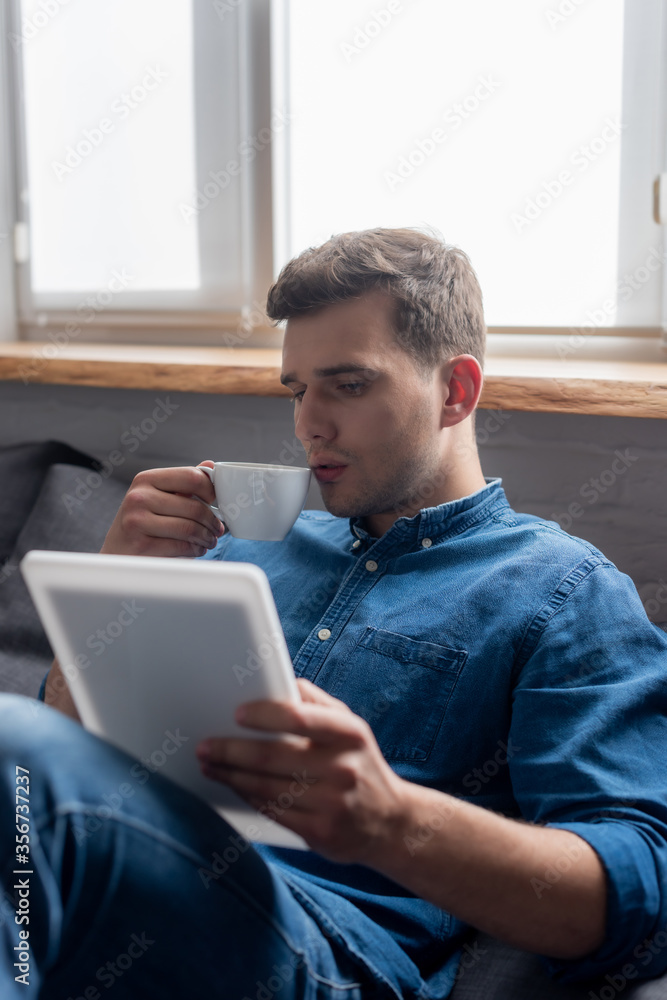 selective focus of handsome man holding cup and looking at digital tablet selective focus of handsome man holding cup and looking at digital tablet