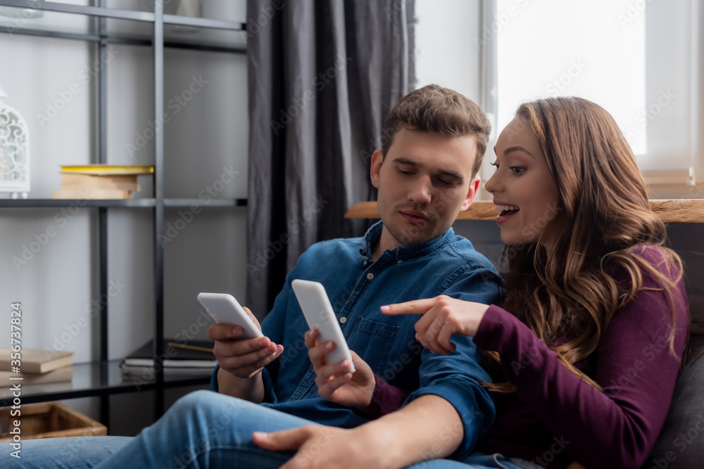 happy girl pointing with finger at smartphone near boyfriend in living room happy girl pointing with finger at smartphone near boyfriend in living room