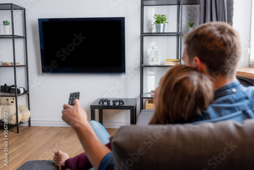 selective focus of man holding remote controller and looking at flat panel tv with blank screen near woman in living room
