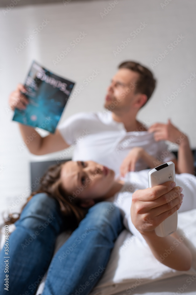 selective focus of girl with remote controller from air conditioner lying near man holding magazine while feeling hot selective focus of girl with remote controller from air conditioner lying near man holding magazine while feeling hot