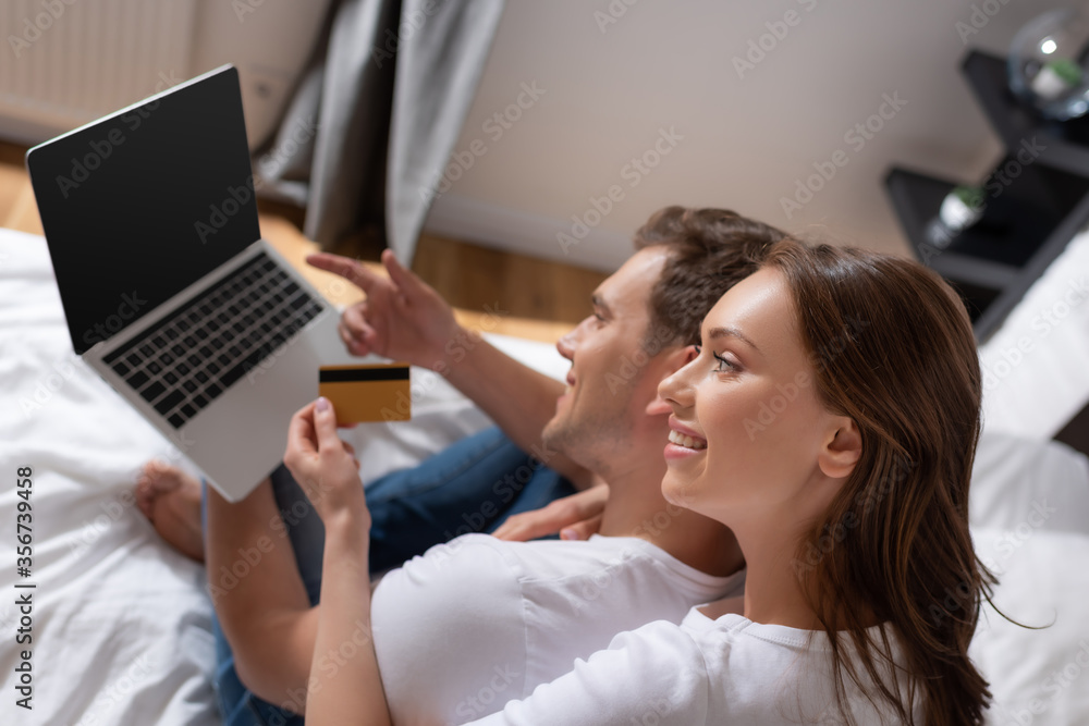Obraz premium selective focus of cheerful woman holding credit card near man using laptop with blank screen in bedroom