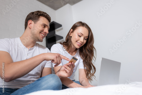 happy man pointing with finger at credit card near laptop and cheerful girl