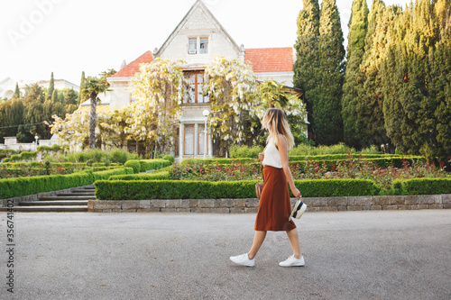 Young beautiful woman, wearing skirt, top and hat, walking in a blooming park among the greenery and the old palace with wisteria. Villa Kharaks (Haraks, Charax), Gaspra, Crimea, Russia. Manor, castle photo